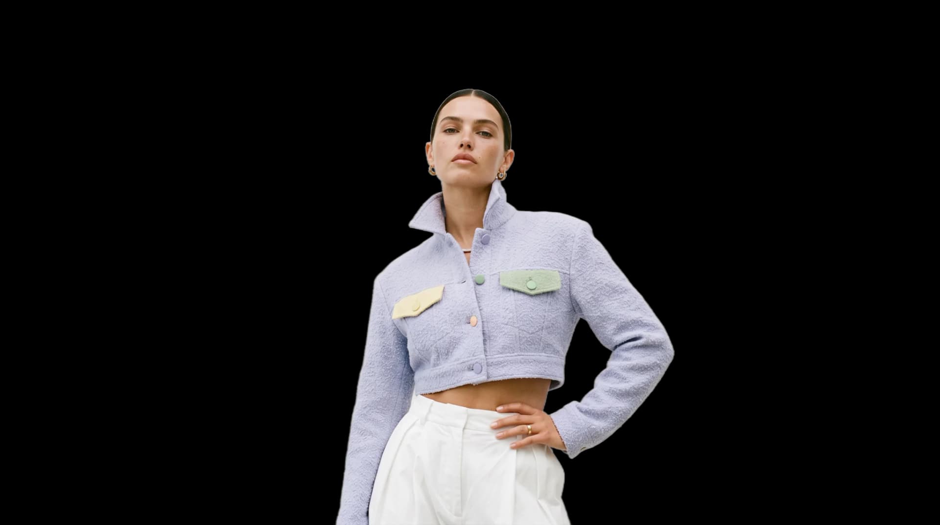 A model in a pale lilac cropped bouclé jacket with pastel pocket flaps and white pleated trousers, one hand on her hip, cut out on a plain ground