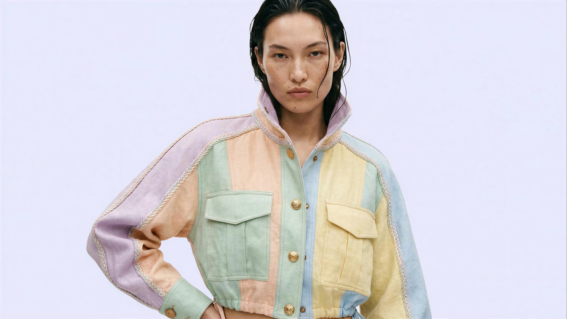 A model in a cropped pastel patchwork linen jacket with gold buttons, on a pale lilac ground