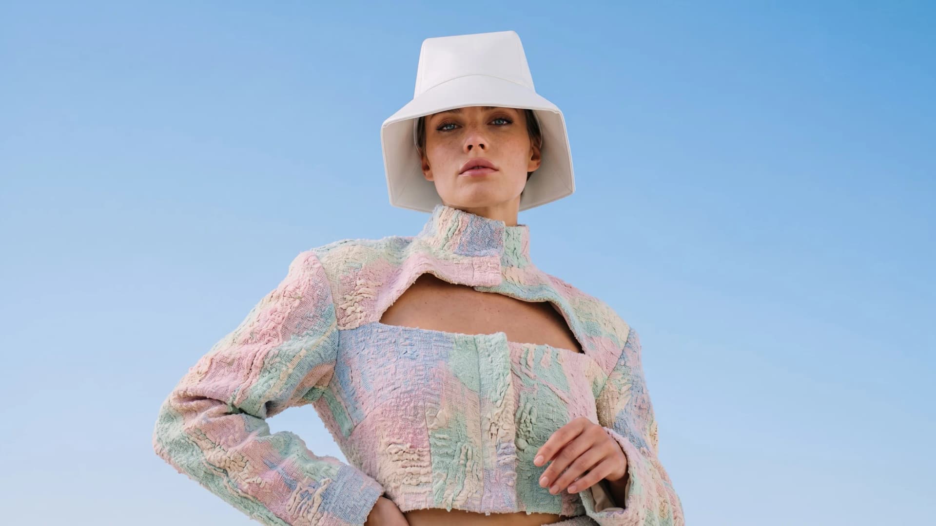Model in a white bucket hat and a cropped pastel patchwork jacket against a blue sky