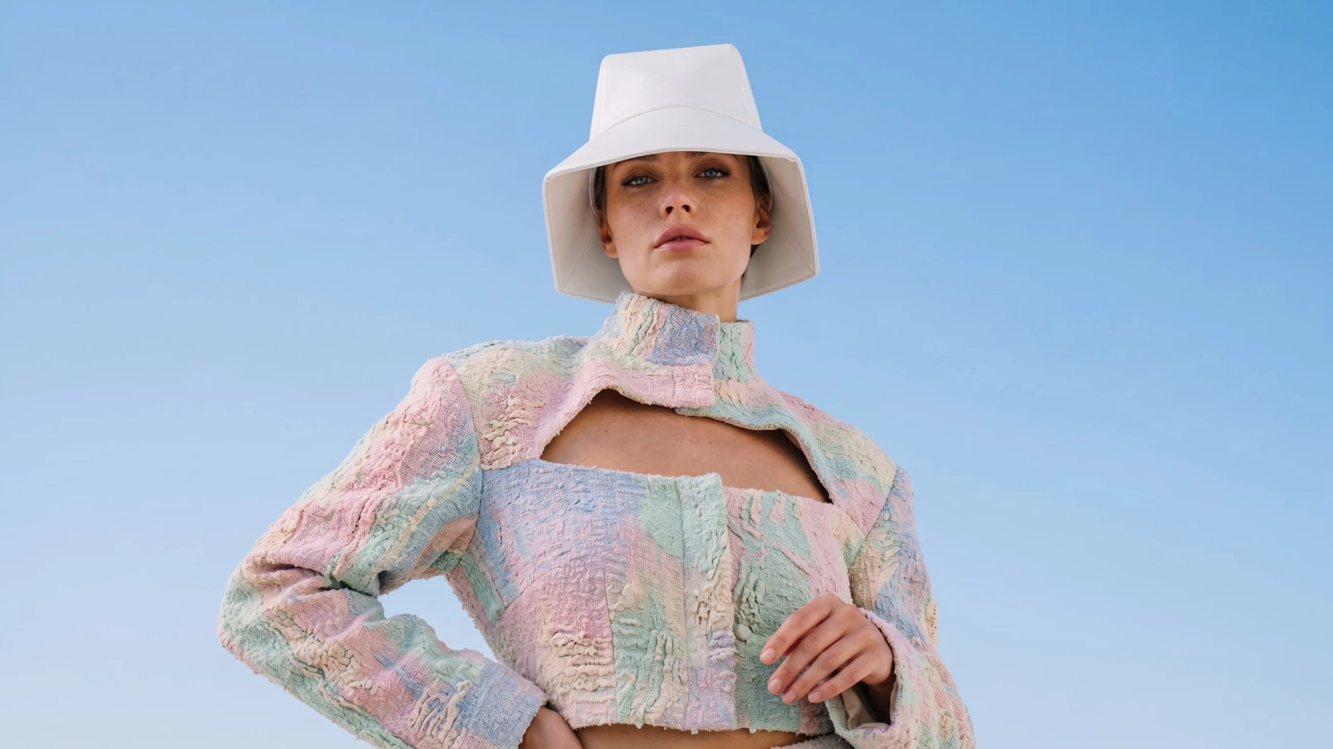 Model in a white bucket hat and a cropped pastel patchwork jacket against a blue sky