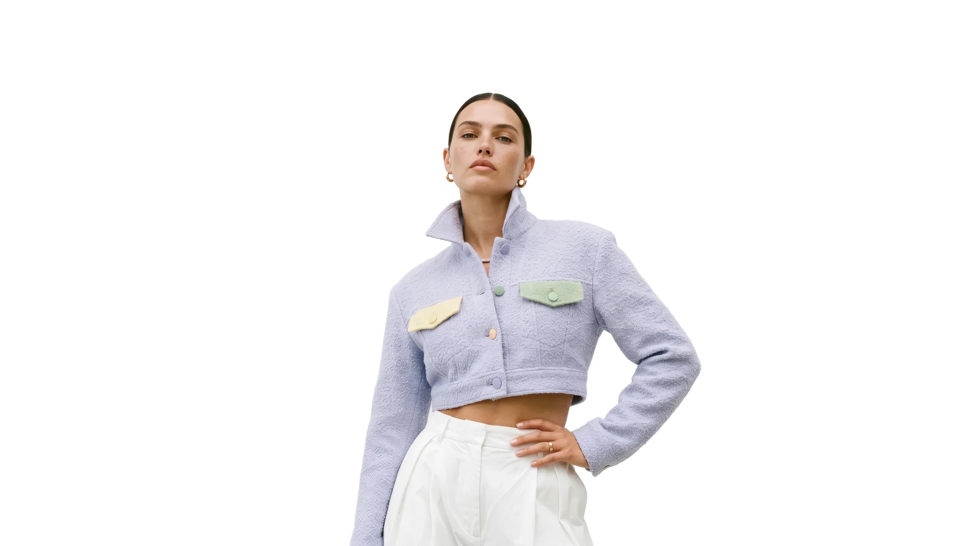 A model in a pale lilac cropped bouclé jacket with pastel pocket flaps and white pleated trousers, one hand on her hip, cut out on a plain ground