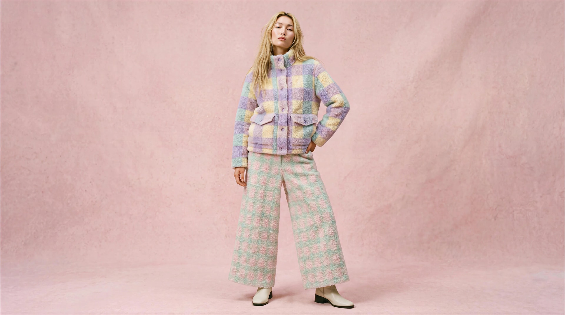 A model in a pastel check jacket and matching wide trousers, standing on a pink studio backdrop