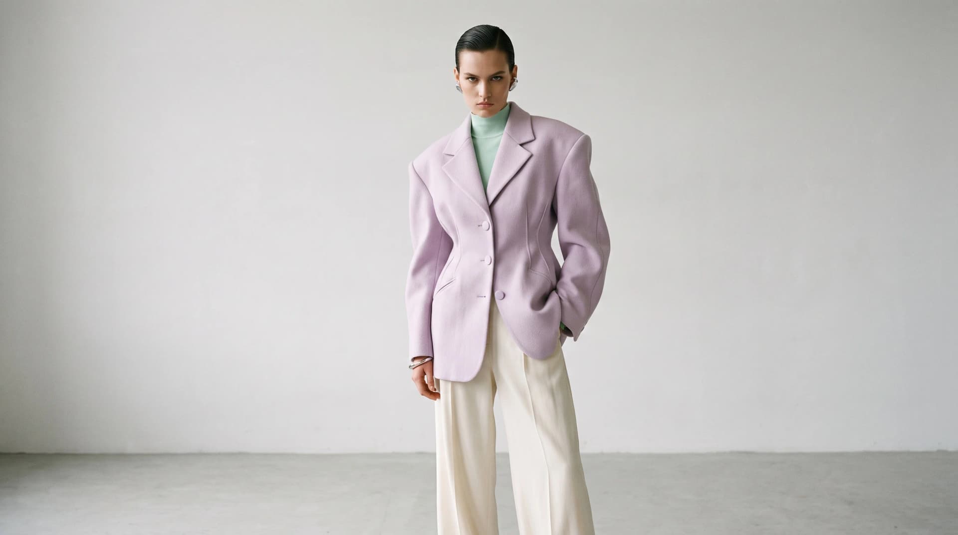 A model in a pale lilac sculptural blazer over a mint high-neck top with cream trousers, standing on a light grey studio ground