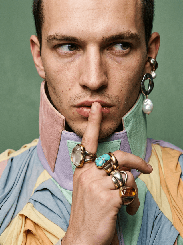 Close-up of a male model with a finger to his lips, pastel leather collar and oversized rings